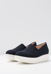 Een paar zwarte instap-platformschoenen met dikke witte gekartelde zolen op een witte ondergrond en een lichtgrijze achtergrond.