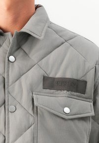 Quilted gray jacket featuring a snap button closure, a textured fabric, and a patch with "REPLAY" on the chest pocket.