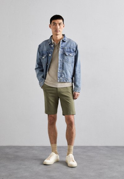 Farmerdzseki, bézs pulóver, olajzöld shorts és fehér edzőcipő. Az öltözet laza szabású, részletes varrással és minimális kiegészítőkkel.