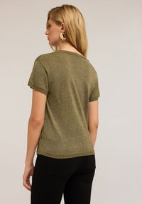 Top de manga corta en verde oliva con un diseño ajustado, acabado metálico texturizado y bordes acanalados, combinado con pantalones negros ajustados.