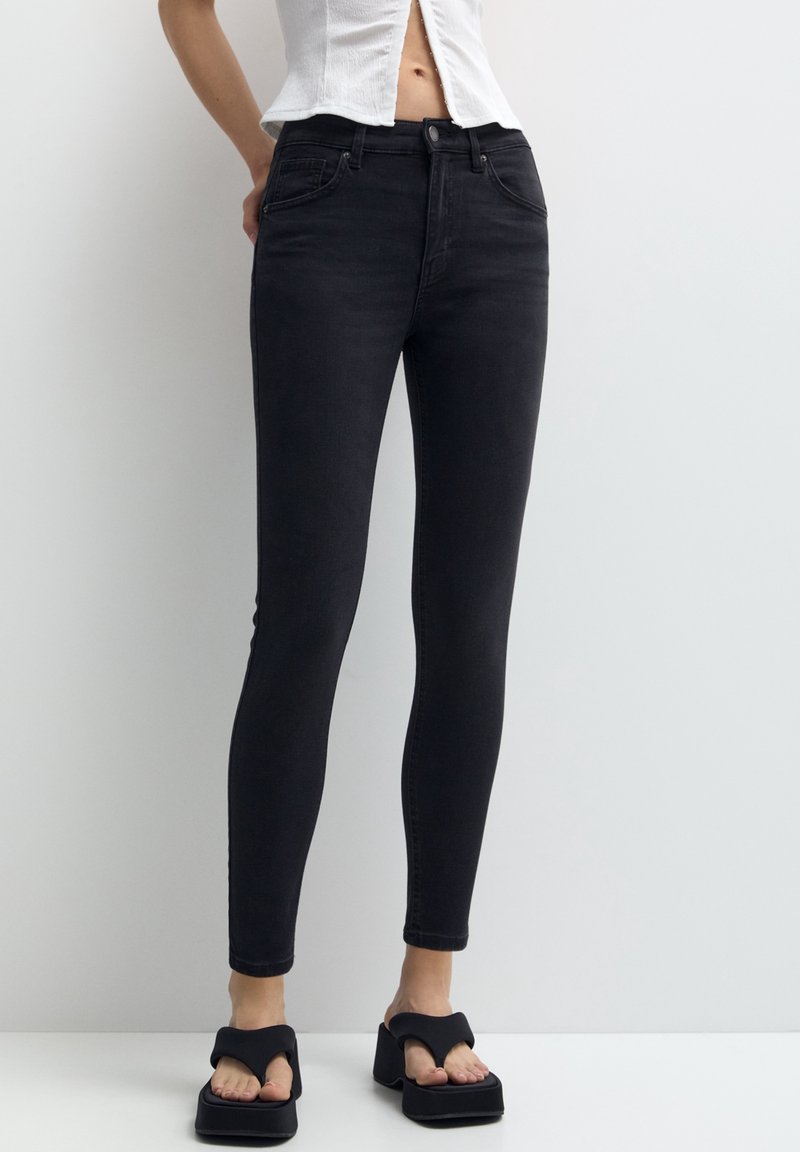 Frau trägt schwarze High-Waist Skinny Jeans, weißes cropped Oberteil mit Knöpfen und schwarze Plateausandalen, steht vor einer weißen Wand.