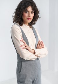 Jasno-beżowa koszula z guzikami pod szarymi ogrodniczkami w wzór rybiej ości, z szerokimi ramiączkami. Modelka ma kręcone włosy i skrzyżowane ramiona, co podkreśla dopasowanie i teksturę stroju.