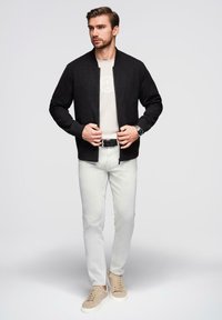 Męska czarna kurtka bombowiec, miękki materiał, zamek z przodu, noszona na jasnobeżowej koszuli z okrągłym logo, w połączeniu z jasnymi spodniami i sneakerami.