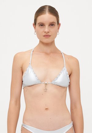 Woman with light skin and brown hair wears a silver metallic bikini with a star pendant on the top, standing against a plain white background.