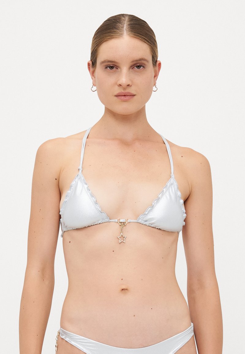 Woman with light skin and brown hair wears a silver metallic bikini with a star pendant on the top, standing against a plain white background.