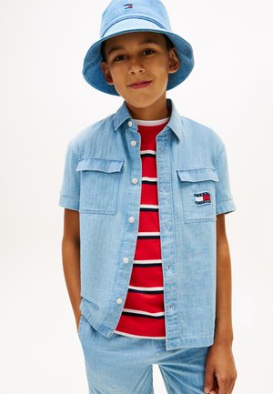 Junge trägt einen blauen Jeans-Bucket-Hut, ein kurzärmeliges Jeanshemd, ein rot gestreiftes T-Shirt und passende Jeanshosen, steht mit den Händen in den Taschen.