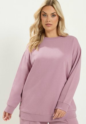 Kobieta o długich blond włosach, ubrana w luźną, długą, mauve bluzkę z długim rękawem i dopasowane spodnie na jednolitym jasnym tle.