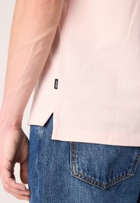 Μαλακή ροζ κοντομάνικη μπλούζα με υφή, πλευρικές σχισμές και μια μικρή μαύρη ετικέτα στο στρίφωμα, συνδυασμένη με μπλε τζιν παντελόνι.