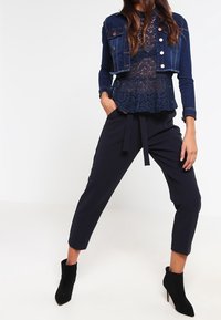 Mörk denimjacka med silverknappar, lager på lager över en marinblå spetsblus. Marinblå skräddarsydda byxor med knytbälte, kombinerat med svarta ankelskor.