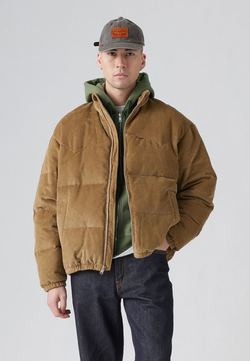 Brun kordfløjs puffjakke med lynlås foran, elastiske manchetter og en afslappet pasform. Båret over en grøn hættetrøje og mørke jeans. Grå kasket på hovedet.