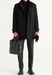 Cappotto nero con colletto alto, chiusura a bottoni e cuciture visibili, abbinato a una borsa nera strutturata e pantaloni grigi a fantasia.