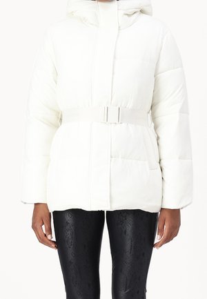 Person trägt eine weiße, mit Gürtel versehene Steppjacke mit Kapuze und schwarze, strukturierte Leggings, steht mit entspannten Armen an den Seiten.