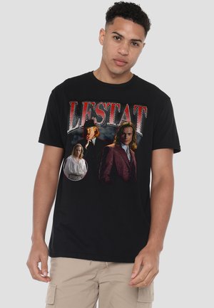 Junger Mann trägt ein schwarzes T-Shirt mit dem Schriftzug "LESTAT" und drei Charakterporträts auf der Vorderseite, kombiniert mit beigen Cargo-Shorts.