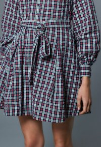 Röd, marinblå och grön rutmönstrad skjortklänning med skärp i midjan, långa puffärmar och veckad kjoldesign. Tygets textur verkar slät.