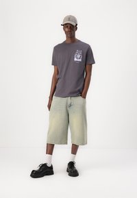 Grå t-shirt med grafisk detalj på bröstet, ljusgula denimshorts, svarta skor med tjocka sulor och en beige keps.