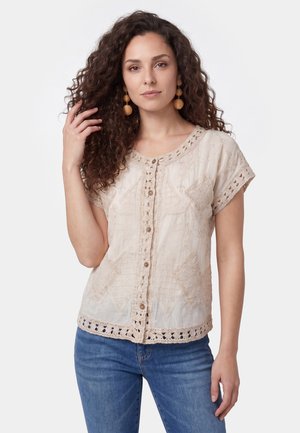 Frau mit lockigem Haar, die große runde Ohrringe trägt und eine beige bestickte kurzärmelige Bluse mit Knöpfen und blauen Jeans.