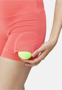 Korallfargede treningstights med en sidelomme som holder en lys grønn tennisball. Stoffet er glat, med flate sømmer og tettsittende passform.