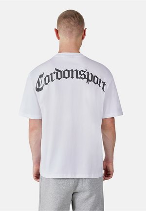 Mann mit kurzen Haaren, der ein weißes T-Shirt mit "Tordonsport" in schwarzer gotischer Schrift auf dem oberen Rücken und hellgraue Shorts trägt.