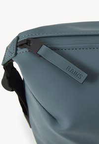 Borsa blu resistente all'acqua con una superficie liscia, chiusura con zip e una tracolla nera regolabile. Etichetta del brand "RAINS" sulla zip.