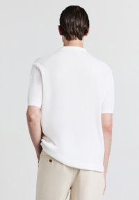 Polo shirt bianco a maniche corte con un motivo testurizzato, colletto e polsini a costine, progettato per un fit rilassato, indossato con pantaloni beige.