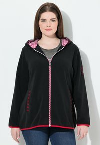 Veste en polaire noire avec une doublure de capuche à carreaux rouge et blanc, zip contrastant, poches latérales avec des accents décoratifs, et une coupe décontractée.