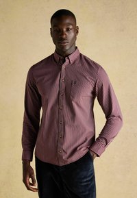 Rød- og hvidstribet skjorte med lange ærmer og knaplukket krave; har en lille logo på brystlommet. Teksturen fremstår glat.