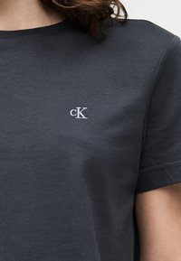 Κοντινό πλάνο ενός ατόμου που φοράει σκούρο γκρι κοντομάνικη μπλούζα με μικρό λευκό λογότυπο "cK" κεντημένο στο στήθος.