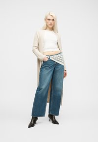 Νεαρή γυναίκα που φορά μακρύ μπεζ ζακέτα, λευκό κοντό τοπ, μπλε τζιν παντελόνι φαρδιάς γραμμής, μαύρα μπότες με τακούνι μέχρι τον αστράγαλο και πλεκτή ζώνη-τσάντα.