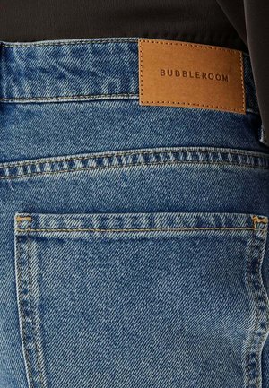 Achteraanzicht van een blauwe spijkerbroek met zichtbare achterzak en een bruine leren patch met het label "Bubbleroom" op de tailleband.