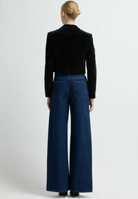 Chaqueta de terciopelo negro con lunares blancos combinada con pantalones vaqueros azules de talle alto y pernera ancha. Textura suave, diseño corto.