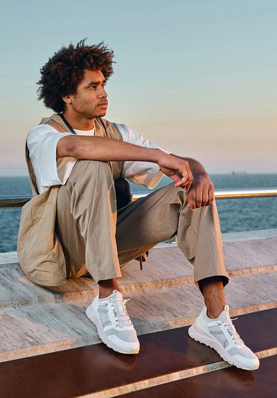 Sneakers atletici bianchi con dettagli in rete, lacci curati e una suola testurizzata. Indossati con abbigliamento beige chiaro in un ambiente costiero.
