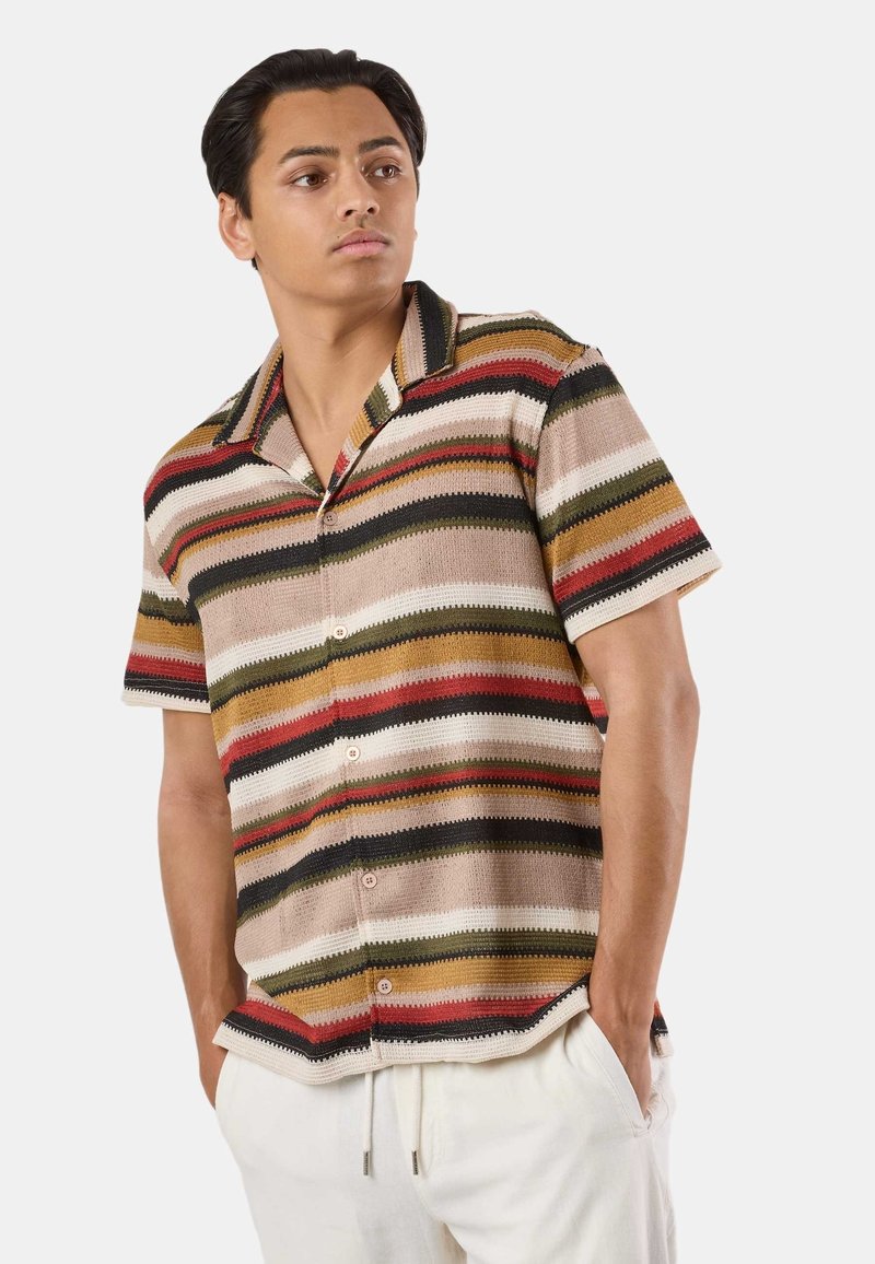 Mladý muž v krátkosrstom tričku s viacfarebnými horizontálnymi pruhmi a bielych nohaviciach, ruky vreckách, pozerá sa bokom.