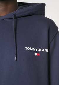 Felpa blu navy realizzata in tessuto morbido, presenta un logo frontale con "TOMMY JEANS" e un emblema colorato. Include un cappuccio con coulisse.