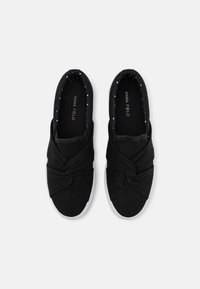 Schwarze Canvas-Slipper mit einem verdrehten Design an der Vorderseite, weißer Gummisohle und einem gepunkteten Muster im Inneren. Markennamen auf der Innensohle sichtbar.