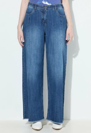 Persoon draagt wijde, twee-kleurige blauwe jeans met rafelige zoom en witte schoenen, staand tegen een lichte achtergrond.