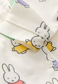Chemise en tissu blanc ornée de lapins dessinanimés vêtus de tenues jaunes et roses. Présente une finition texturée avec une fermeture à boutons et un col plié.