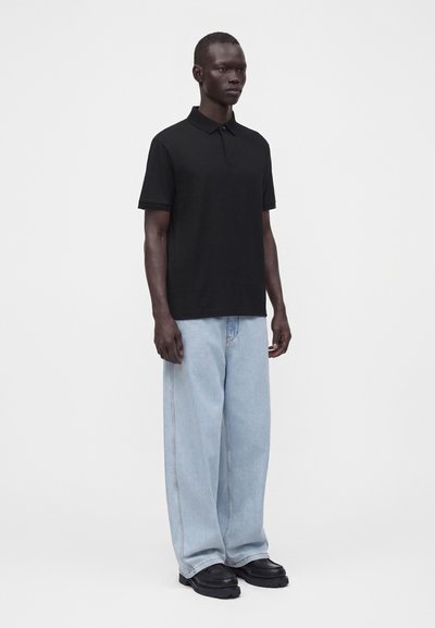 Homme adulte portant un polo noir à manches courtes, un jean bleu clair à jambes larges et des chaussures noires, debout devant un fond uni.
