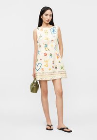 Femeie care poartă o rochie albă fără mâneci, cu imprimeuri florale abstracte colorate, ținând o geantă de mână verde măsliniu și purtând sandale negre.