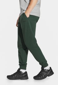 Zielone spodnie dresowe z kieszeniami i zwężanym krojem, zestawione z czarnymi butami sportowymi o teksturowanej powierzchni i białymi skarpetkami.