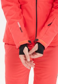 Helle rote Jacke aus wasserdichtem Material mit verstellbaren Manschetten, featuring ein schwarzes Innenfutter und Klettverschluss.