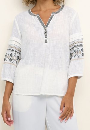 Kvinde iført en hvid tekstureret bluse med sort og beige geometrisk broderi på ærmer og halsudskæring, kombineret med hvide bukser.