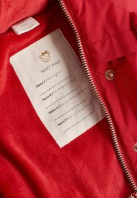 Veste bébé rouge avec zip ouvert révélant une étiquette avec une icône d'ours, la marque NEXT baby, et des espaces pour écrire quatre prénoms de bébé et leurs descriptions.