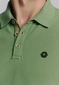 Groene polo met een gestructureerde stof, een sluiting met drie knopen en decoratieve knopen, en een hexagonaal logo-patch.