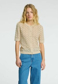 Beige pletený cardigan s diamantovým krajkovým vzorem, kulatým výstřihem, krátkými rukávy a zvlněným lemem; má přední zapínání na knoflíky.