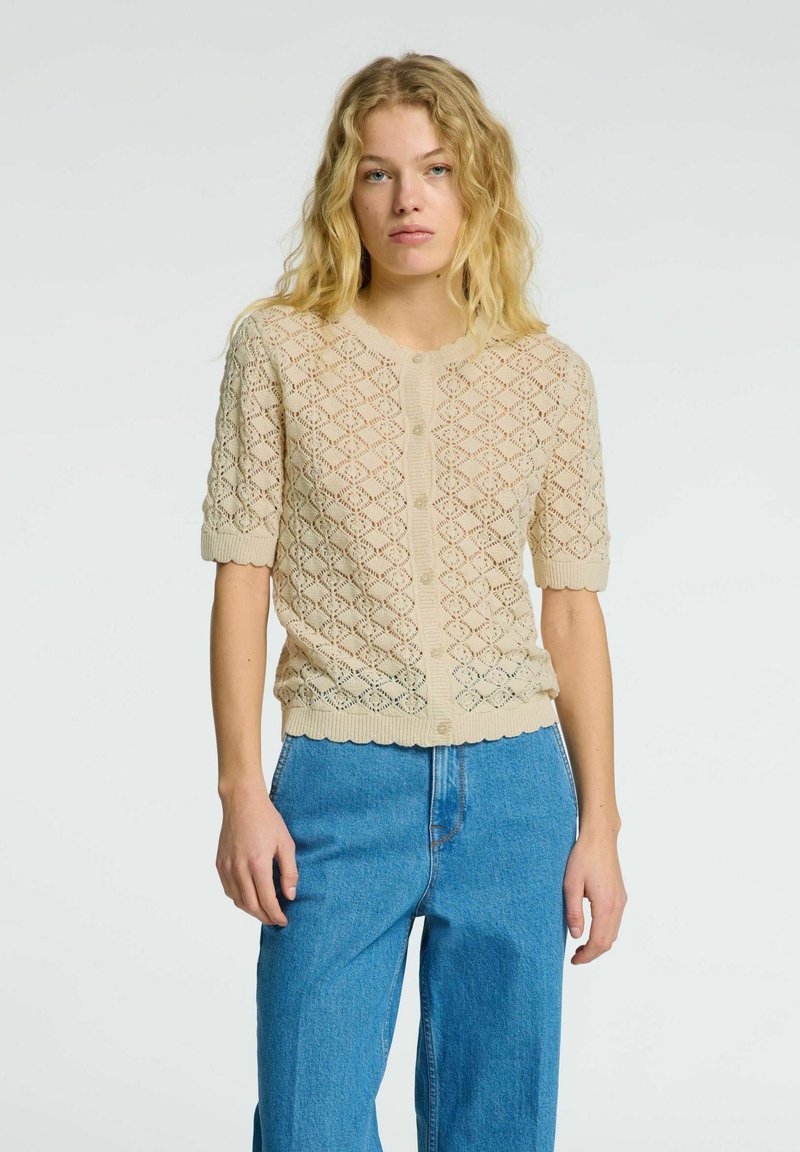 Beige pletený cardigan s diamantovým krajkovým vzorem, kulatým výstřihem, krátkými rukávy a zvlněným lemem; má přední zapínání na knoflíky.