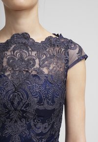 Merenan sininen mekko, jossa on yksityiskohtainen pitsi päällyste kimalteilla, aaltoileva pääntie ja lyhyet olkaimet. Silkkinen satiinikangas.