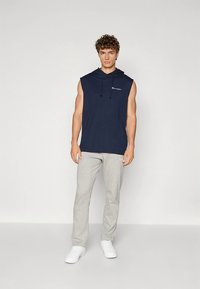 Mornarska brezrokavnik s kapuco z logotipom spredaj, kombiniran z svetlo sivimi športnimi hlačami in belimi supergami. Material je videti mehak in udoben.