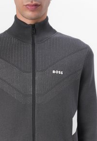 Grau gestrickte Zip-Jacke mit hohem Kragen. Texturierte Einsätze und ein Logo auf der Brust. Sie verfügt über ein figurbetontes Design und athletische Stilakzente.