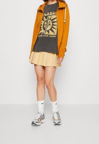 Felpa arancione con zip, t-shirt grigia con grafica, gonna pieghettata beige, calzini sportivi bianchi e scarpe da corsa argento con dettagli arancioni.