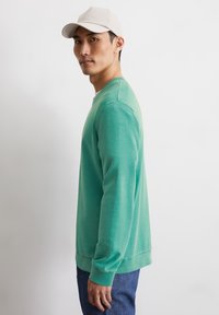 Zielony sweatshirt wykonany z bawełny, z długimi rękawami, okrągłym dekoltem i ściągaczami. Nosi się go z niebieskimi dżinsami, na jasnym tle.